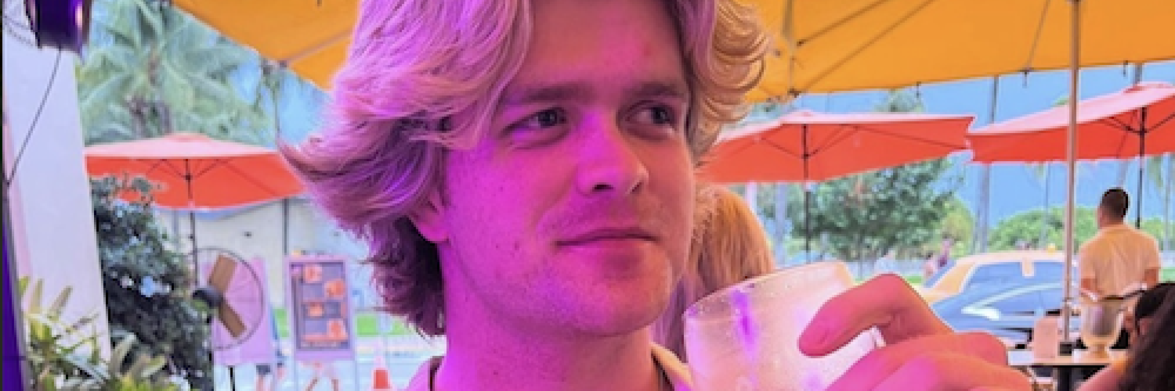 Matt sipping a white wine with table umbrellas behind him. The image hue has been manipulated with some hues of pink.